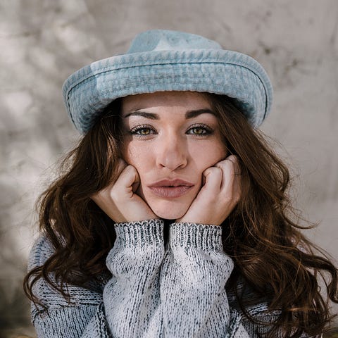 Close-up of woman with long, dark hair. She’s wears a round-brimmed hat and grey sweater. Her hands, with fingers turned in, rest on her chin.