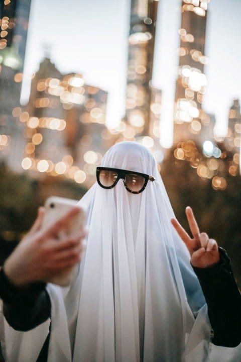 A foto de uma pessoa com um lençol branco cobrindo todo o corpo, utilizando um óculos escuro e tirando uma selfie com seu smartphone, fazendo o sinal de paz e amor com a outra mão. Ao fundo, uma cidade desfocada à luz do dia.