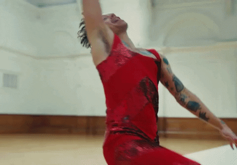 Imagem GIF do artista Harry Styles, do seu clipe de As It Was. Ele veste um macacão vermelho brilhante e faz movimentos descontraídos de dança, tanto em um ambiente interno como externo.