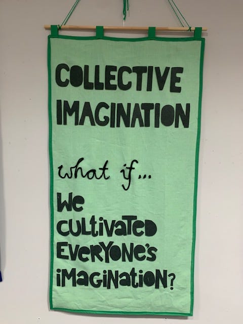 Showing a green canvas banner with the writing ‘collective imagination what if we culutvated everyone’s imagination’ stiched on in black fabric. Banner created by Time Rebels Ruth and Odette of We Are Makers