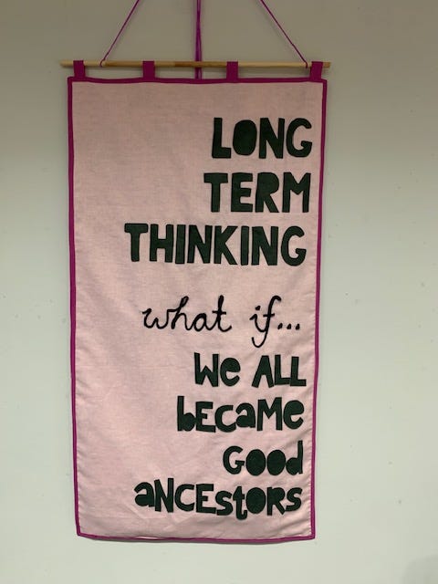 Showing a pink canvas banner with the writing ‘long term thinking what if we all become good ancestors?’ stitched on in black fabric. Banner created by Time Rebels Ruth and Odette of We Are Makers