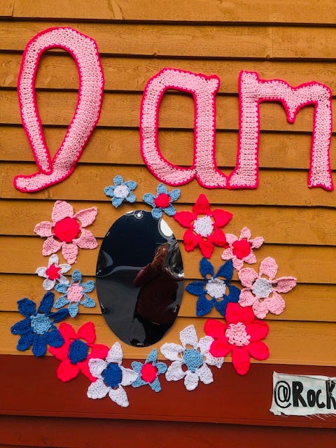 An outdoor wall festooned with crocheted “I am”, colourful flowers and a mirror.