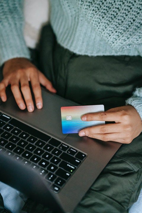 A lady wearing a primarily green outfit, holds a credit card between her lap and her laptop, online payment activity is assumed.