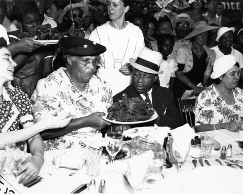 A black and white photo of an integrated group of people having a banquet. At the center, a Black couple sits, and the woman holds a plate of turkey towards the man.