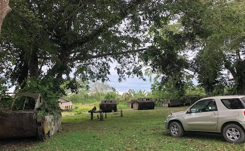 A small SUV car next to four abandoned World War II amphibious vehicles in a field on a tropical island