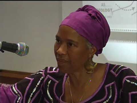 Marimba Ani, mulher negra usando turbante e blusa roxas, falando ao microfone.