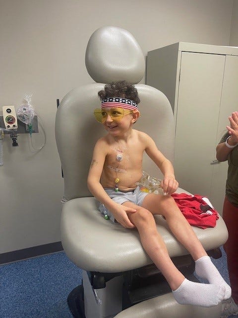 A young boy with a bandana on his head is seated in a chair in a medical setting, appearing calm and at ease.