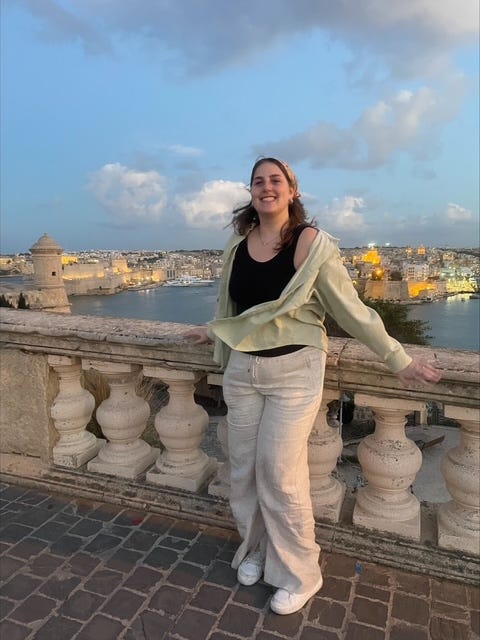 A girl wearing a white pants, a black shirt tank top and a light green overshirt stands on a bridge overlooking a city landscape.