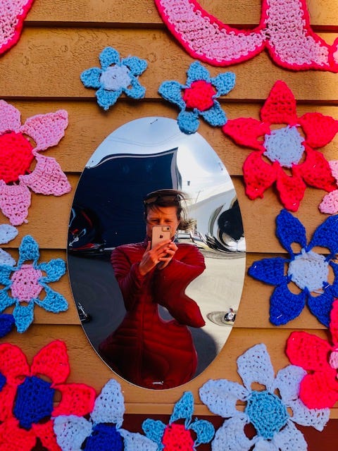 Nina Elliott takes a photo of herself in a distorted mirror surrounded by colourful crocheted flowers hung on an outside wall.