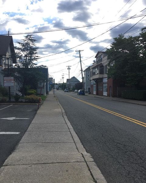 A long road two-lane road with a double yellow stripe down the middle a sidewalk to the left and buildings on either side. No traffic.