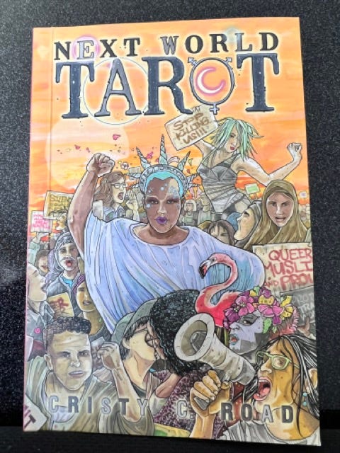 A picture of the front of a tarot deck called “Next World Tarot” with a series of protestors at a rally, featuring a black woman dressed as the Statue of Liberty.