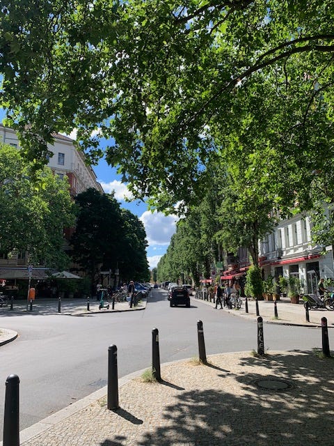 Photo of a treelined street in Berlin. It’s quiet with only few people and one car. The sky is a beautiful blue and the sun is shining.