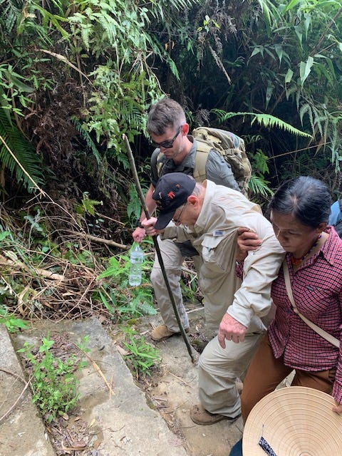 An elderly man hikes through the jungle with people on either side of him, holding him up.