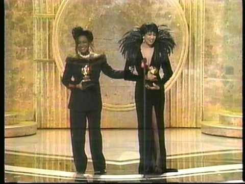 Patti LaBelle clasps Lisa Fischer’s right elbow with her left hand as they stand against a columned gold and marble background while holding their Grammy awards after a tie win. In this moment, Patti has ceded the perched microphone to Lisa to finish giving her acceptance speech to the audience.