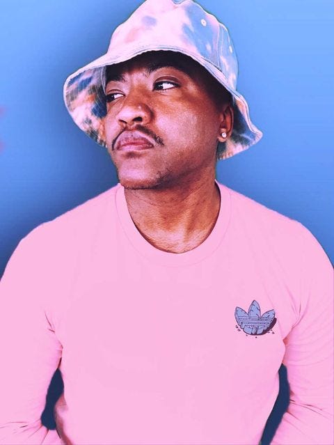 Kai looks off to the left in a pink shirt and patterned bucket hat. He’s in front of a blue background.