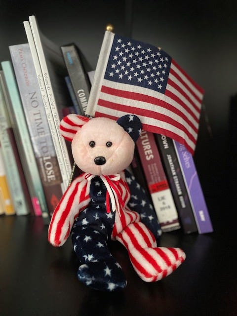 A red, white, and blue beanie baby sitting on a bookshelf with an American flag behind it.