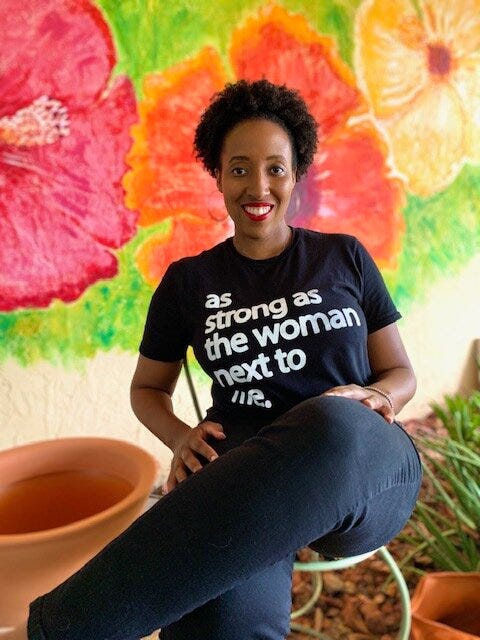 Black Haitian-American womxn with short cropped hair, sitting in a black shirt that says “as strong as the woman next to me” against a floral background.