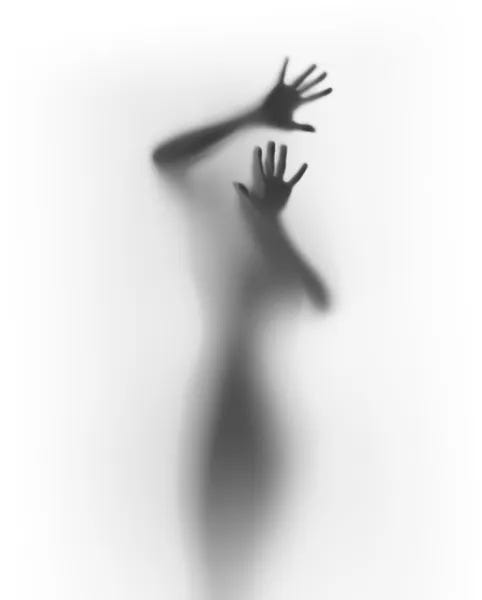 A photograph of an individual, with their hand pressed against the surface, is struggling to break free from the foggy white glass. Most of the photo is just a white background with just the trapped individual in the center. The individual’s face is unseen and the dark silhouette of the rest of their body can only be seen in the photo.