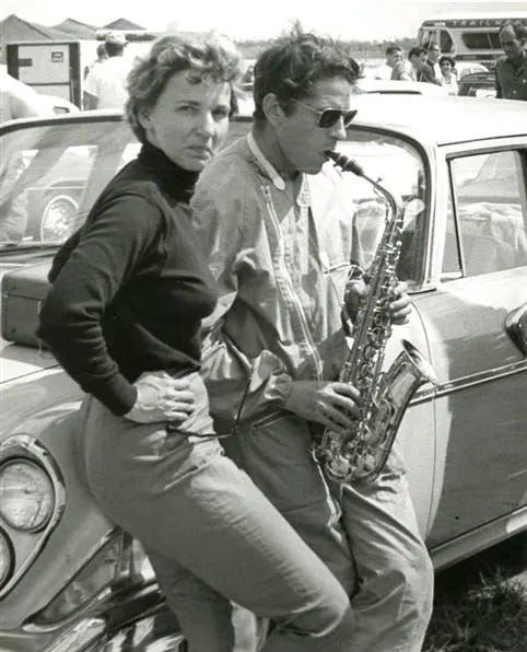 In a black-and-white photo, a woman in a black turtleneck with her hands on her hips looks directly at the camera. Next to her, a man wearing sunglasses plays the saxophone. Both lean against the hub of a car.