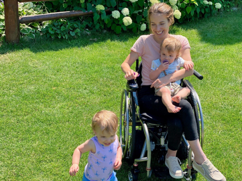 Dani Izzie in her wheelchair, legs crossed, holding one twin on her lap, another twin running by the side of the wheelchair. They are outside on the grass on a sunny day