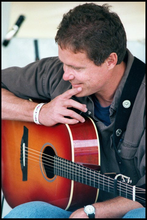 Photograph of singer-songwriter Dave Carter resting on his guitar, which is resting on his knee, as he listens to another songwriter perform a song at a workshop stage at the Falcon Ridge Folk Festival on 29 July 2000, two years before he died. Photo copyright 2023 by Patrick T. Power. All rights reserved.