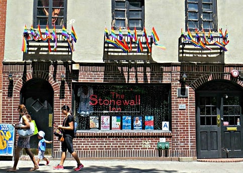 A brick building adorned with pride flags at the top. There’s a red neon sign that reads “The Stonewall Inn”. A family walks by the building.