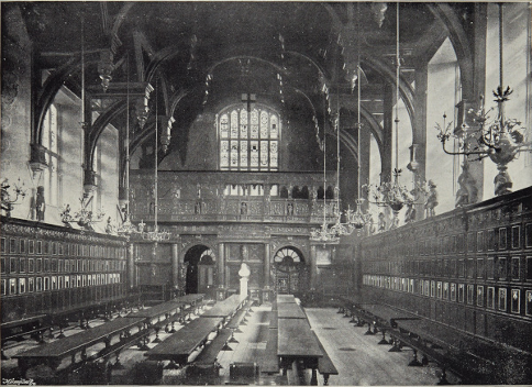 A black and white photograph of a large hall with long tables and benches and very high ceilings and stained-glass arched windows