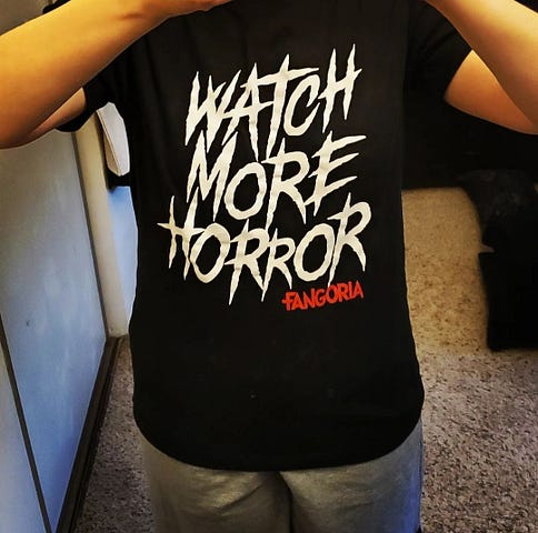 Black t-shirt with the phrase “Watch More Horror” in white font and “Fangoria” in red font.