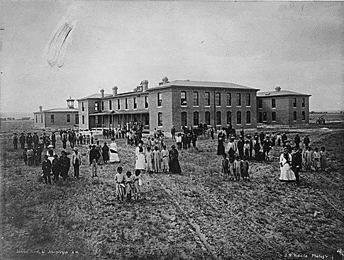 A black and white photo depicts a large school building with surrounding structures. Groups of adults and children scattered around the school grounds all face in the direction of the camera.