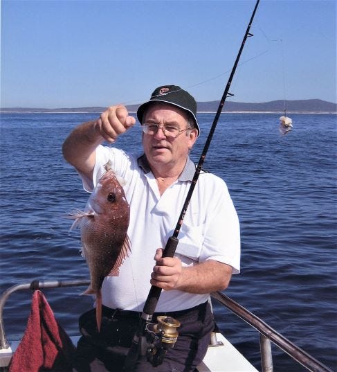 A man in a white polo shirt and navy blue bucket hat holds a fishing rod and a caught fish aloft. He has a gently pleased expression.