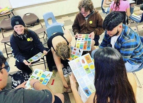 A group of young people dressed casually and sitting in classroom chairs, playing a game. They are all holding sheets of paper with different boxes and images on them.