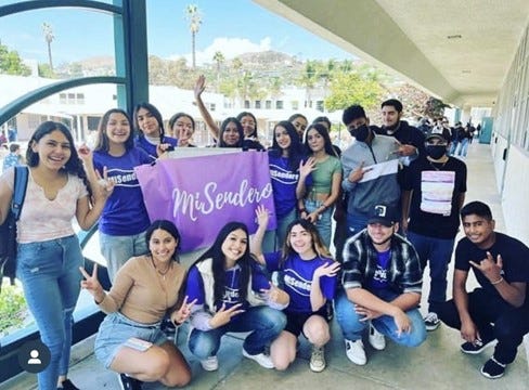 A group of students on a high-school campus holding a purple flag that says “MiSendero.” A lot of them are wearing blue shirts with the same logo, and they are all smiling with their arms lifted. It is a sunny day in the background.