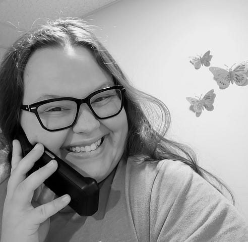 Allison, the author, holding a flip phone to her face and smiling. She has dark long hair, fair skin, and glasses. The photo is in black and white. Butterfly art hangs on the wall behind her.