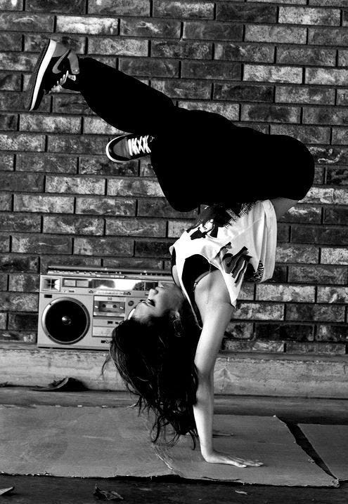 A black and white picture of a bgirl in a freeze upside down in the street in front of a brick wall.