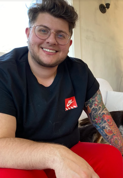 Me smiling on a white couch. You can see my beard has grown in, my hairline has changed, and my jawline is much more defined. I also now have a tattoo representing my transition.