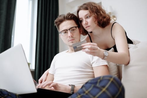 FOTO DE CASAL OLHANDO AS CONTAS FINANCEIRAS COM CARA DE SURPRESA SENDO QUE ELA SEGURA O CARTÃO DE CRÉDITO E ELE O NOTEBOOK.