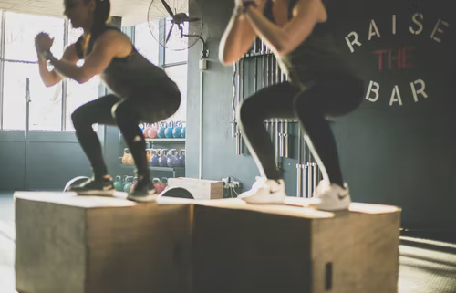 Two women performing box jumps together at a gym