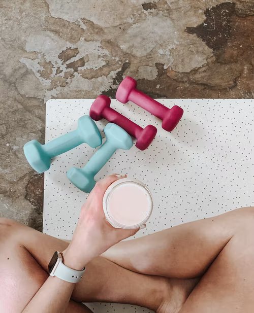 Woman sitting on the floor with a pre workout drink in hand and blue and burgundy dumbbells in front