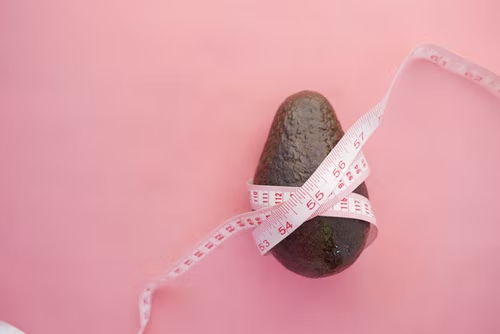 An image of an avocado wrapped with an inch tape on a pink background.