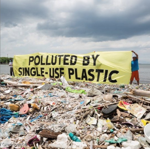 A pile of plastic waste polluting a beach area with two individuals holding a sign “polluted by single-use plastic.”