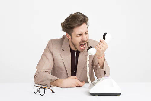 A frustrated man screaming into a telephone with his glasses on the table