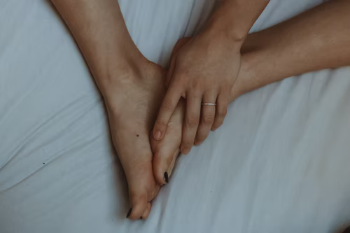 A woman’s hand holding her feet, sitting over a white bedsheet.