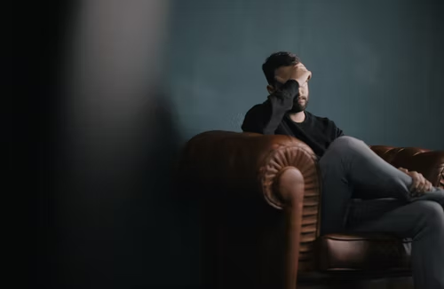 A man sitting on a sofa which his hand on his head.