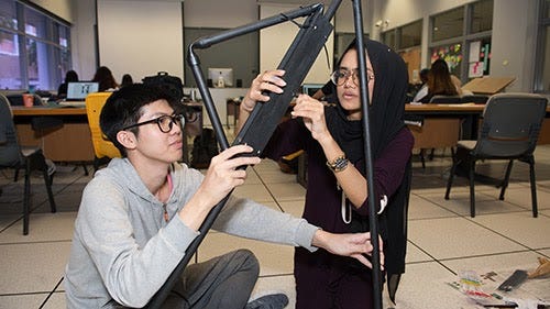 A man wearing glasses and a woman wearing glasses and a headscarf work together to build a prototype with black pipes.