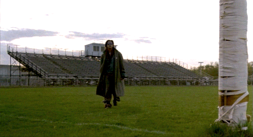 A person in a trench coat walks across a sports field, approaching the camera. As they get closer, they raise their fist in victory.