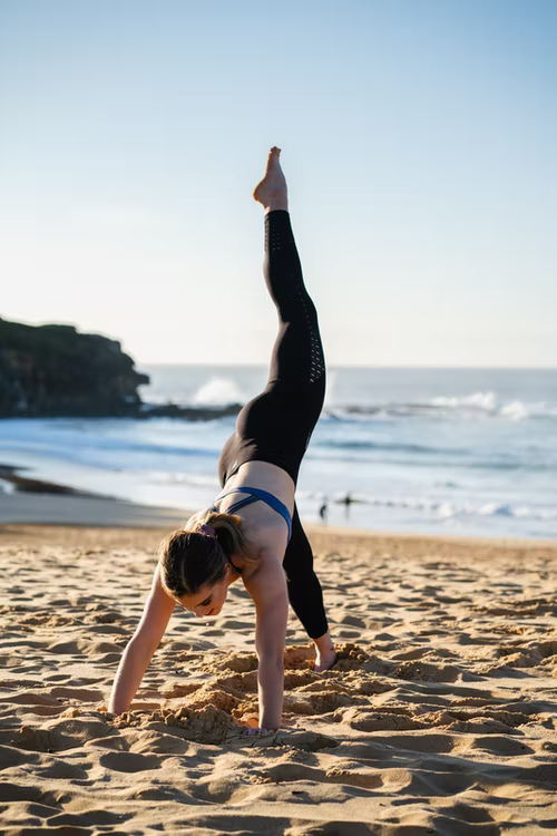 A woman doing a yoga pose on a beach with the ocean behind her, wearing black leggings with one leg straight in the air and her hands on the ground