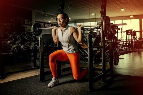 A woman in orange leggings performing lunges with a barbell in a squat rack at a gym