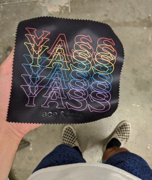 A glasses cloth laid out in someone’s palm. The design reads ‘YASS’ written repeatedly in different rainbow colours.