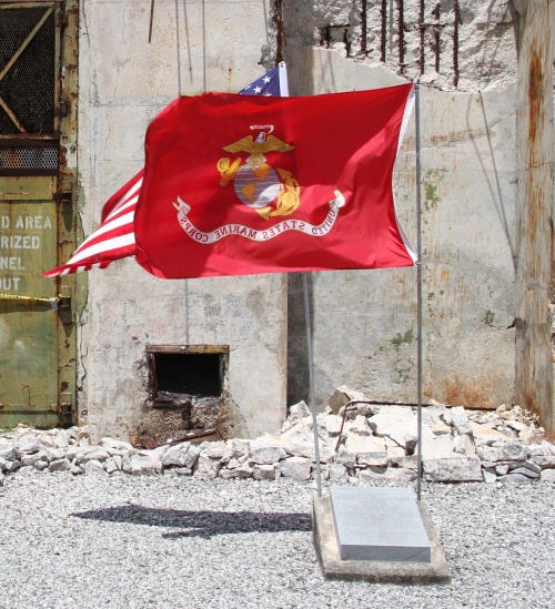 The Marine Corps flag is mounted above a memorial stone dedicated to Lt. George Cannon. It sits in front of a building that appears to be falling apart.