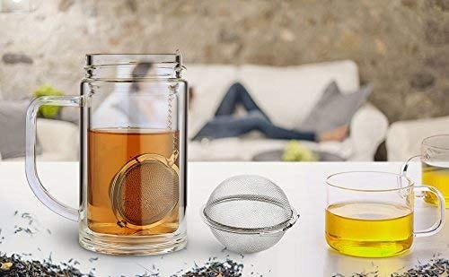 A glass stein full of black tea sits on the left side of the image, a metal tea ball sitting within the liquid. It’s chain is hooked over the top lip of the glass. To the right of that is an empty tea ball. To the right of that is a smaller glass mug with green tea inside. In the background, the blurry form of a person can be seen stretched out on a couch.